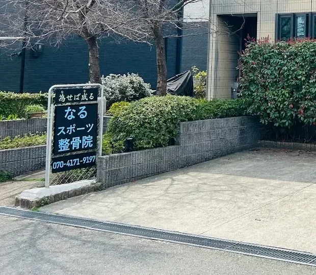 なるスポーツ整骨院の外観。2階建ての建物の前に「為せば成る なるスポーツ整骨院」と書かれた看板が立ち、手前に駐車スペースがある。